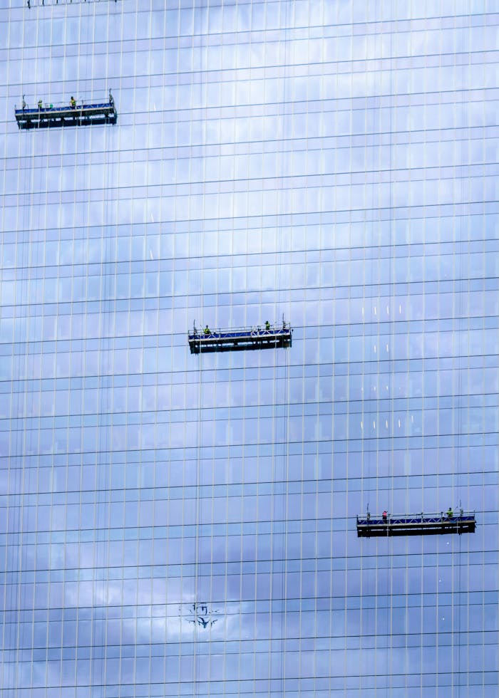 Workers on suspended platforms cleaning skyscraper windows under a cloudy sky.